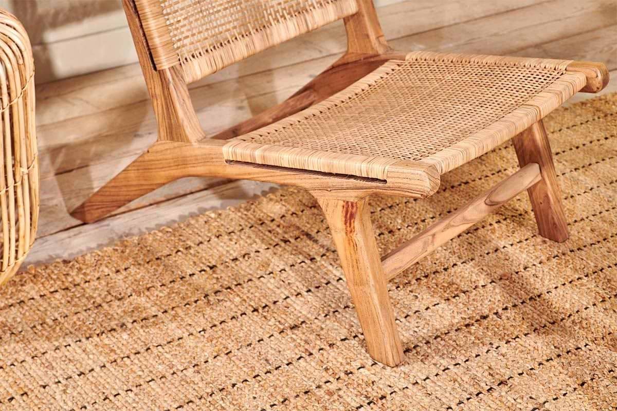 A close-up of the nkuku Madrisana Acacia & Rattan Woven Chair sits on a textured natural fiber rug. Warm, neutral tones and sunlight cast soft shadows on the light wood floor, enhancing the cozy minimalist vibe.
