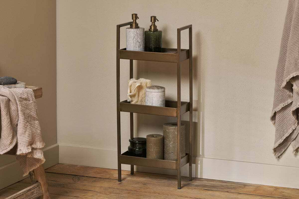 The nkuku Mahi Bathroom Storage Caddy - Antique Brass sits elegantly in a corner, offering stylish three-tiered storage for hand soap, lotion, rolled towels, and candles. Its antique brass finish complements the wooden floor and beige wall, while textured towels hang nearby on a rack and a wooden surface holds more folded towels.