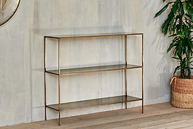 The nkuku Mahi Console Table, a minimalist gold metal and glass shelving unit, stands against a light gray wall on a wooden floor. A leafy green plant in a woven basket sits to the right, with part of a round mirror visible on the left.