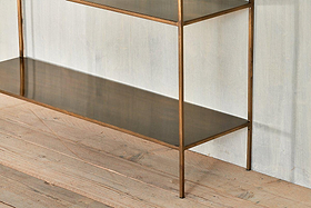 A close-up of the nkuku Mahi Console Table, a minimalist metal design with elegant shelves and a bronze-toned frame, standing on a light wooden floor against a textured wall. Its modern look features clean lines and reflective surfaces.