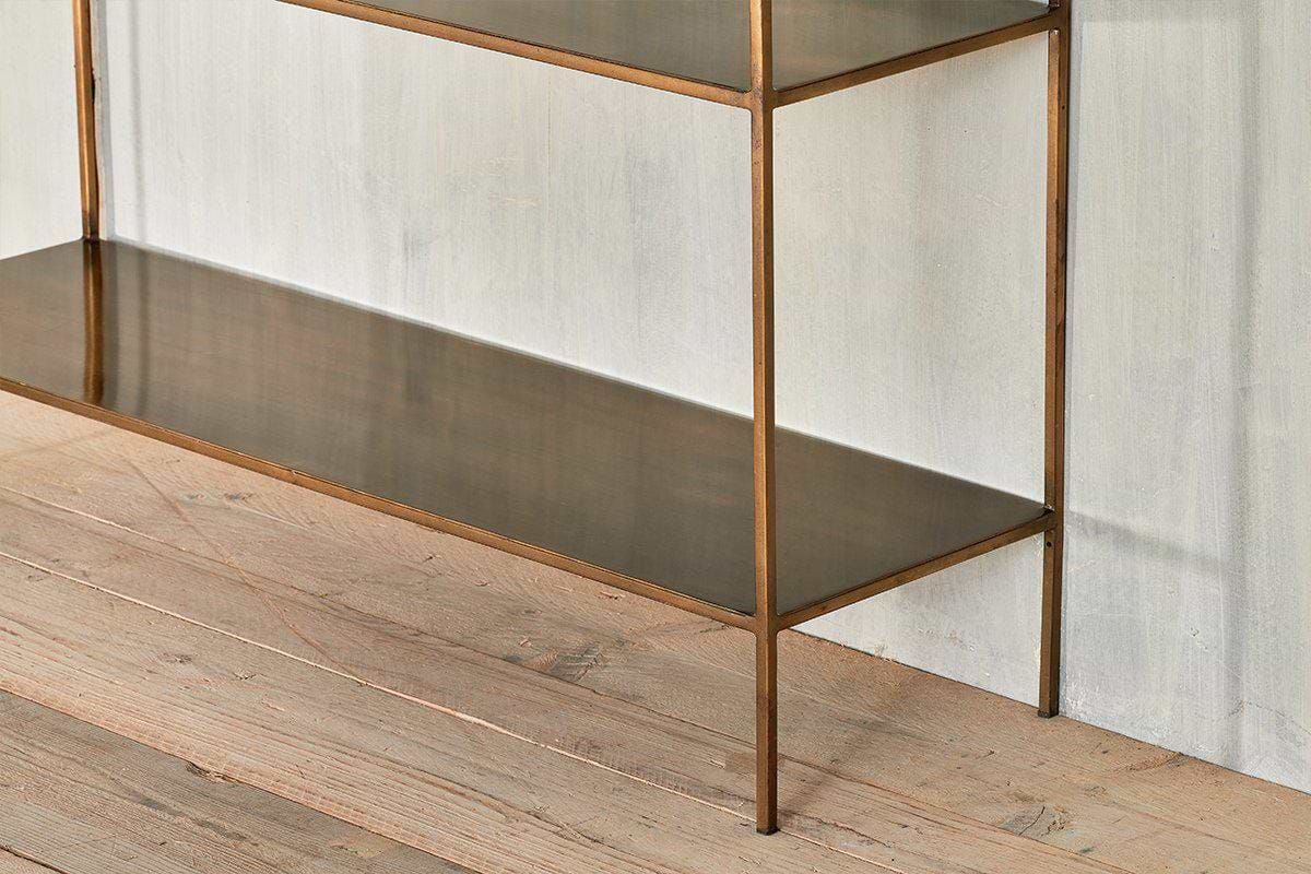 A close-up of the nkuku Mahi Console Table, a minimalist metal design with elegant shelves and a bronze-toned frame, standing on a light wooden floor against a textured wall. Its modern look features clean lines and reflective surfaces.