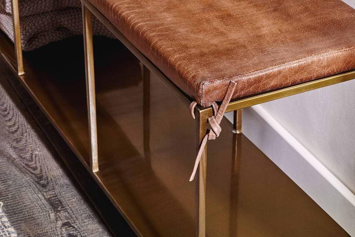 A close-up of the nkuku Mahi Iron & Leather Bench shows a brown leather cushion with thin ties, a brass frame, lower metal shelf, dark wood flooring, and a textured rug partially visible on the left.
