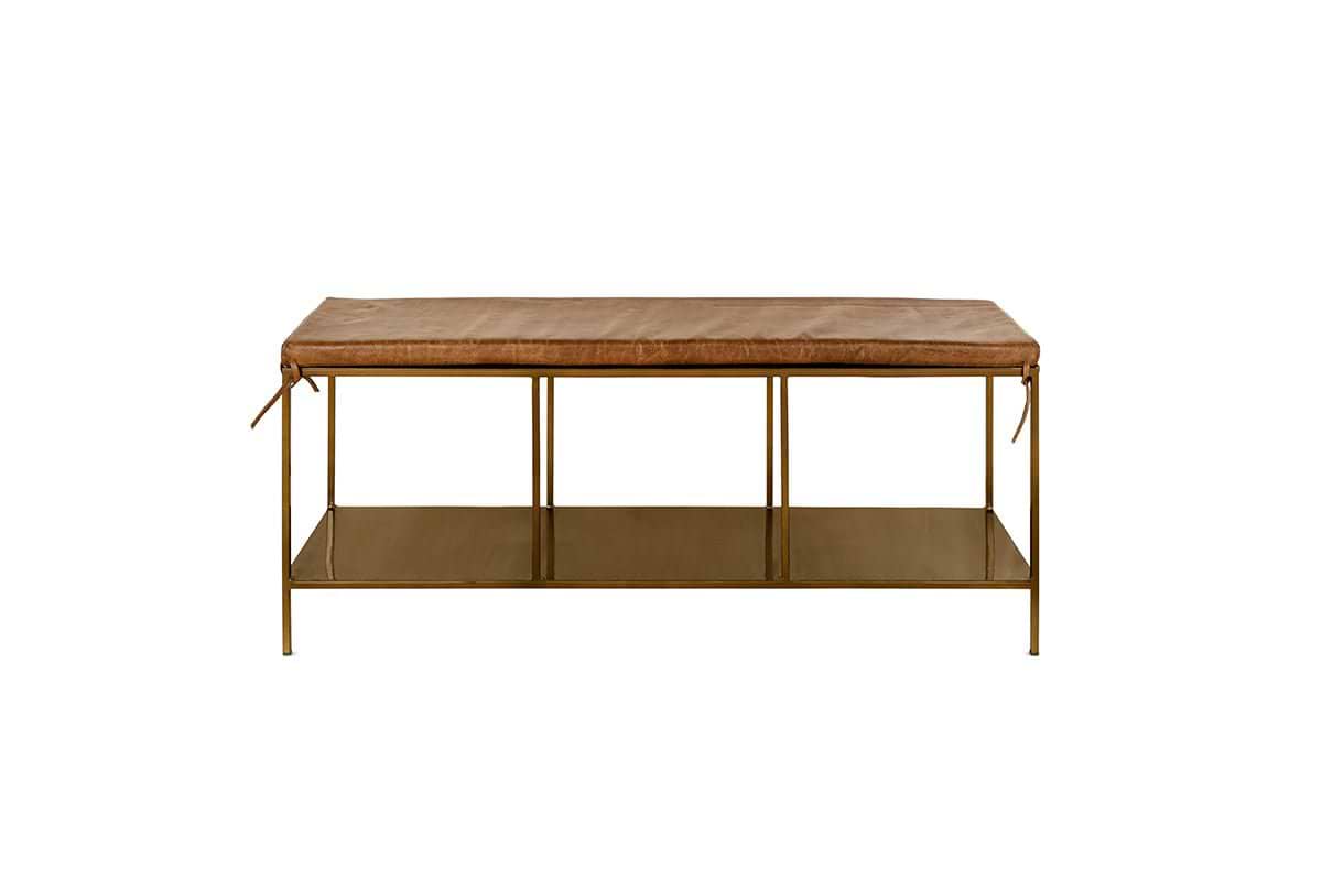 The nkuku Mahi Iron & Leather Bench features a rectangular tan leather cushion with side ties, slender brass legs, and a metal lower shelf. This minimalist bench is photographed against a plain white background.