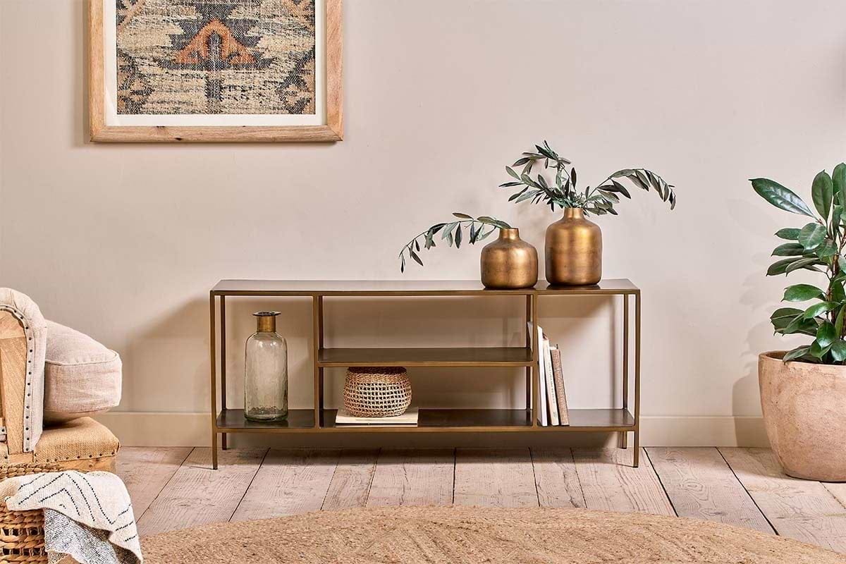The nkuku Mahi Media Standing Shelf, a modern gold metal console with brass shelves, displays books and décor beside a beige armchair and potted plant on light wood flooring. A framed textile artwork hangs above.