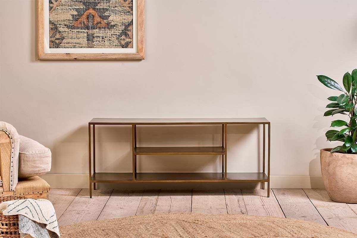 The nkuku Mahi Media Standing Shelf, with gold metal and brass shelves, stands against a beige wall. A potted plant is on the right, a cream armchair with a throw on the left, and a geometric tapestry hangs above on the wooden floor.