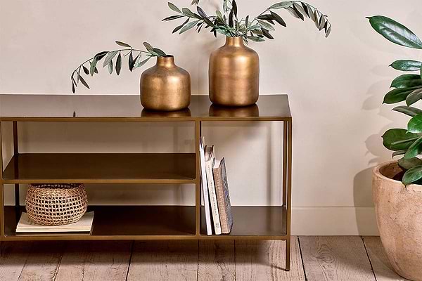 The Mahi Media Standing Shelf by nkuku, featuring a bronze finish, displays two gold vases with leafy branches, books, and a small woven basket. To the right on the wooden floor sits a potted green plant against a cream-colored wall.