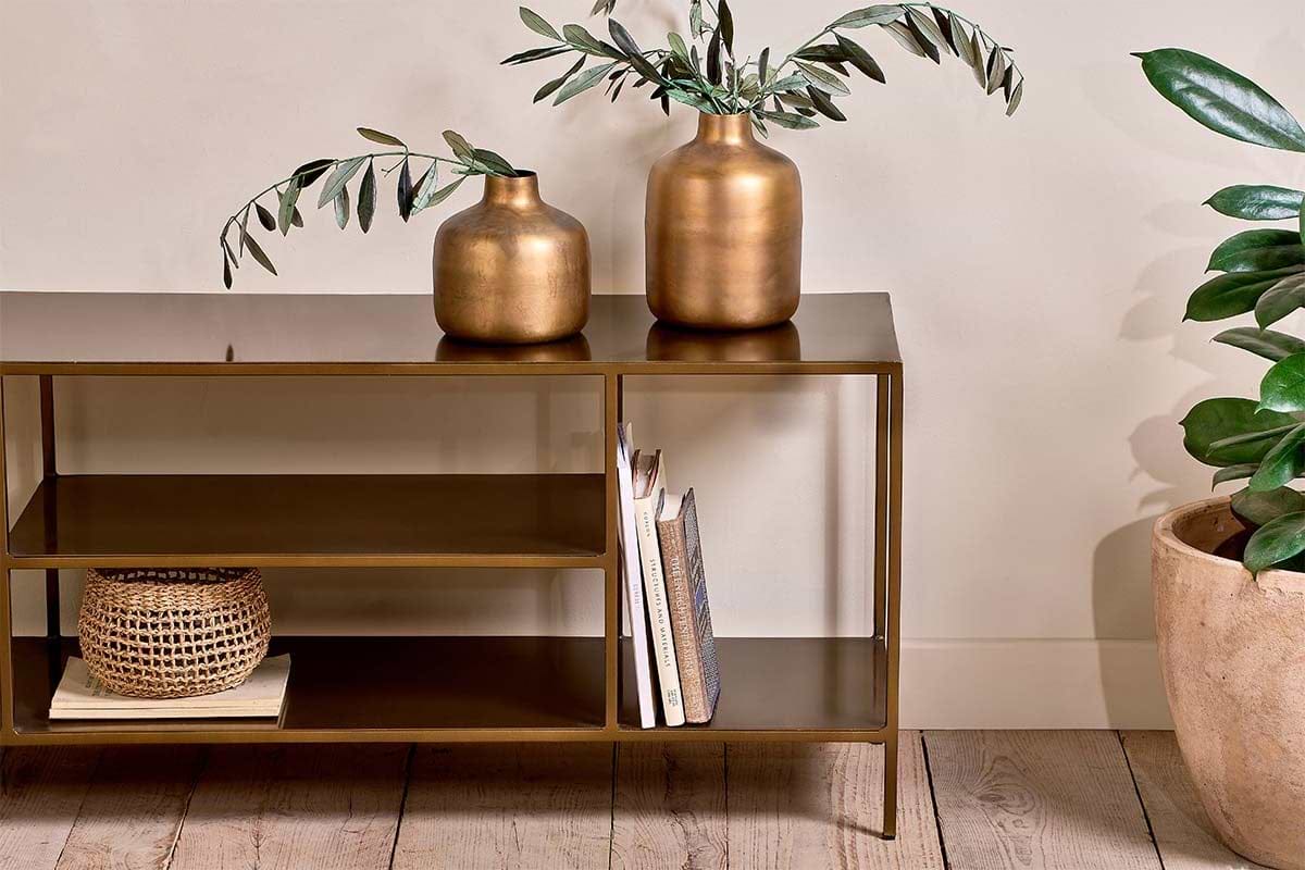 The Mahi Media Standing Shelf by nkuku, featuring a bronze finish, displays two gold vases with leafy branches, books, and a small woven basket. To the right on the wooden floor sits a potted green plant against a cream-colored wall.