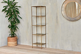 The Mahi Shelving Unit - Narrow by nkuku, featuring four brass shelves, stands beside a large potted plant in a woven basket. A round decorative mirror hangs on the textured wall behind, creating a stylish storage display.