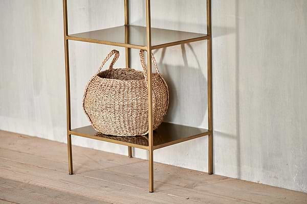 A woven basket with two handles sits on the bottom shelf of the nkuku Mahi Shelving Unit - Narrow, a minimalist brass piece placed against a light textured wall and wooden floor for stylish storage display.