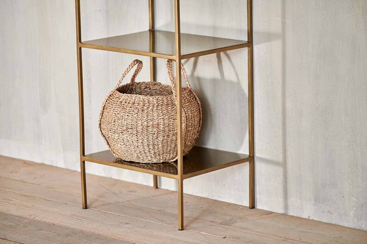 A woven basket with two handles sits on the bottom shelf of the nkuku Mahi Shelving Unit - Narrow, a minimalist brass piece placed against a light textured wall and wooden floor for stylish storage display.