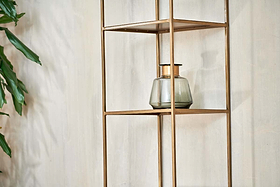 A glass jar with a dark lid rests on the nkuku Mahi Shelving Unit - Narrow, its sleek brass shelves contrasting against a light wall. A leafy green plant peeks in from the left, enhancing the minimalist and contemporary vibe.