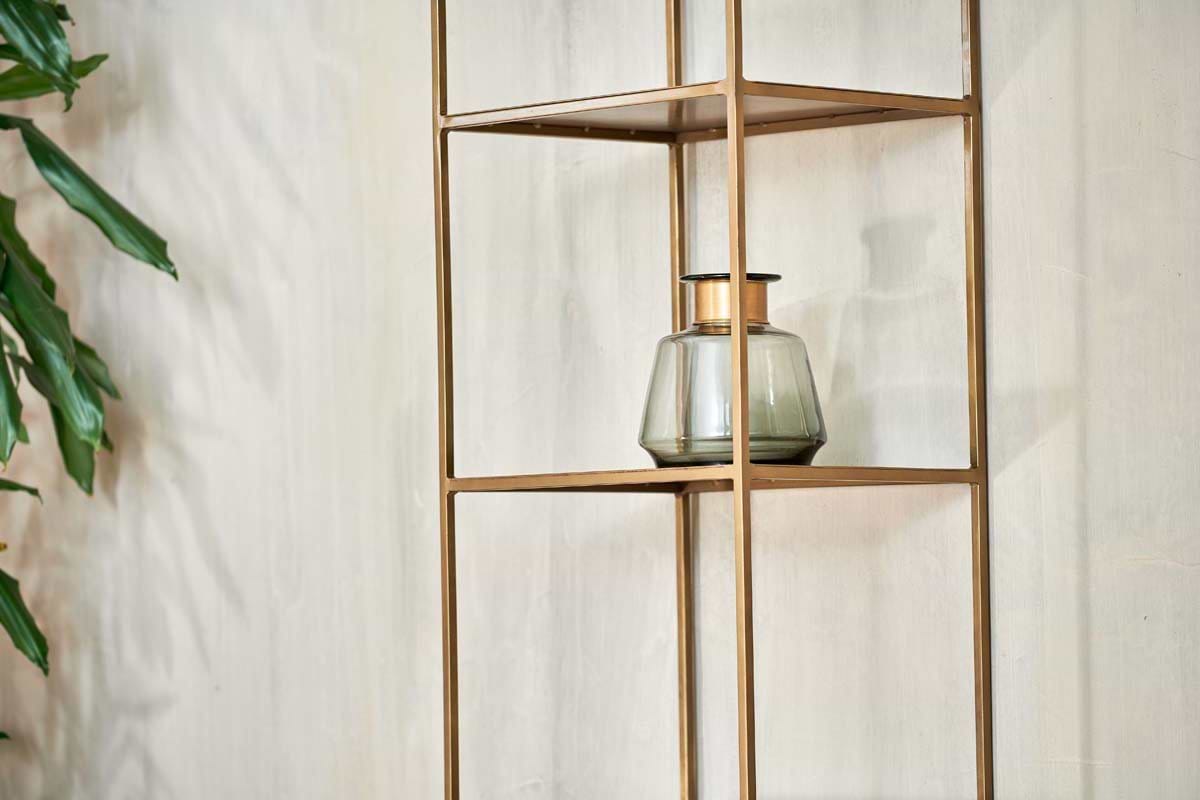 A glass jar with a dark lid rests on the nkuku Mahi Shelving Unit - Narrow, its sleek brass shelves contrasting against a light wall. A leafy green plant peeks in from the left, enhancing the minimalist and contemporary vibe.