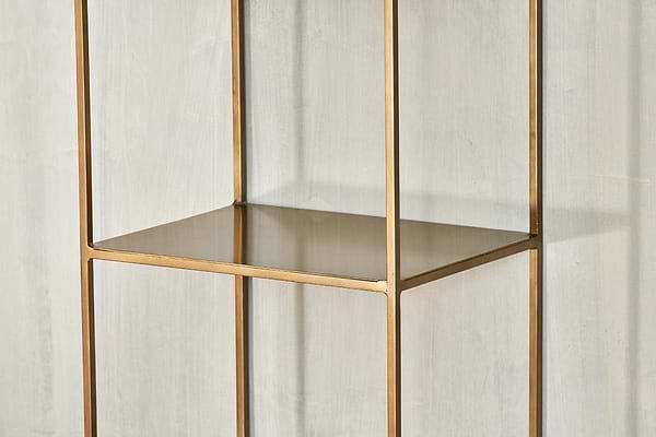 A close-up of the empty, gold metal Mahi Shelving Unit - Narrow by nkuku, featuring a square frame and open design, placed against a light gray textured wall. This brass shelf offers minimalist lines and modern, practical storage.