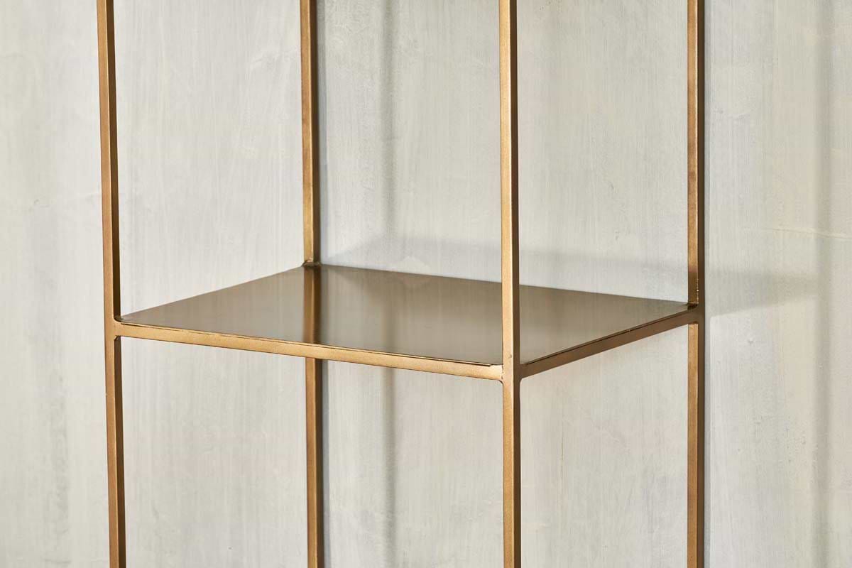 A close-up of the empty, gold metal Mahi Shelving Unit - Narrow by nkuku, featuring a square frame and open design, placed against a light gray textured wall. This brass shelf offers minimalist lines and modern, practical storage.