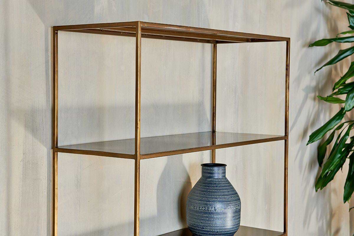 A minimalist nkuku Mahi Shelving Unit - Wide with a brass frame stands against a beige textured wall. On the middle shelf is a dark blue patterned ceramic vase, and a leafy green plant is partially visible on the right.