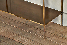 A close-up of the nkuku Mahi Shelving Unit - Wide, showcasing brass shelves with slim legs and a flat surface. Set against a light wall on rustic wooden floorboards, it's ideal for stylish storage displays.