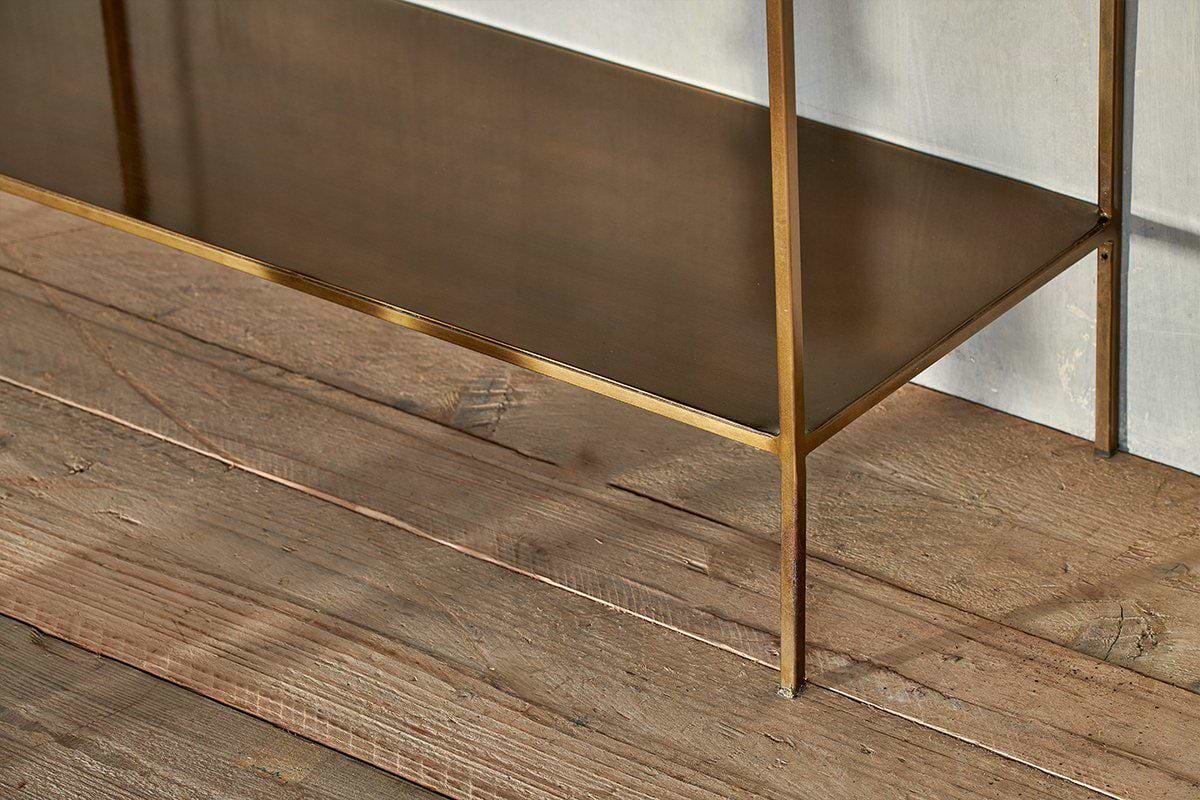 A close-up of the nkuku Mahi Shelving Unit - Wide, showcasing brass shelves with slim legs and a flat surface. Set against a light wall on rustic wooden floorboards, it's ideal for stylish storage displays.