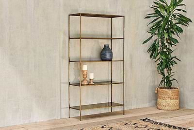 The nkuku Mahi Shelving Unit - Wide, featuring four brass and glass shelves, displays a blue vase, wood candlesticks, and a ceramic cup against a beige wall. A tall leafy plant in a basket stands nearby on the light wood floor.