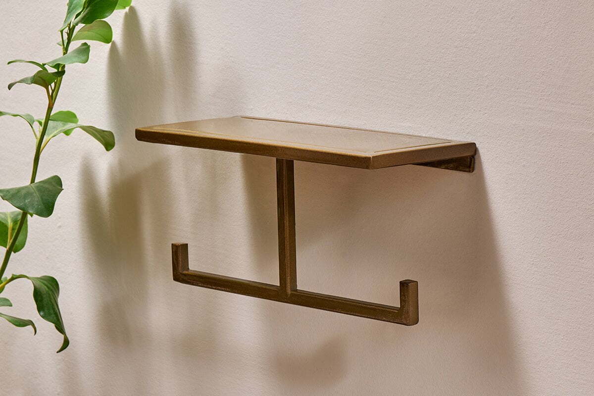 The Mahi Wall Mounted Toilet Roll Holder by nkuku, a small rectangular brass piece with a flat top and two-hook bar, is fixed to a beige wall; a green leafy plant appears partially on the left side.