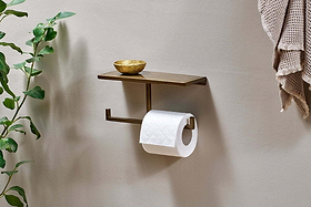 The nkuku Mahi Wall Mounted Toilet Roll Holder in metal holds a gold dish above the toilet roll. A leafy plant is on the left, and textured beige towels hang on the right against a light wall.