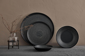 The nkuku Mahika Tray in black, featuring a dark, hand-etched ceramic texture, is styled on a gray fabric surface against a beige wall. A clear glass vase with bare branches completes this minimalistic and muted setting.