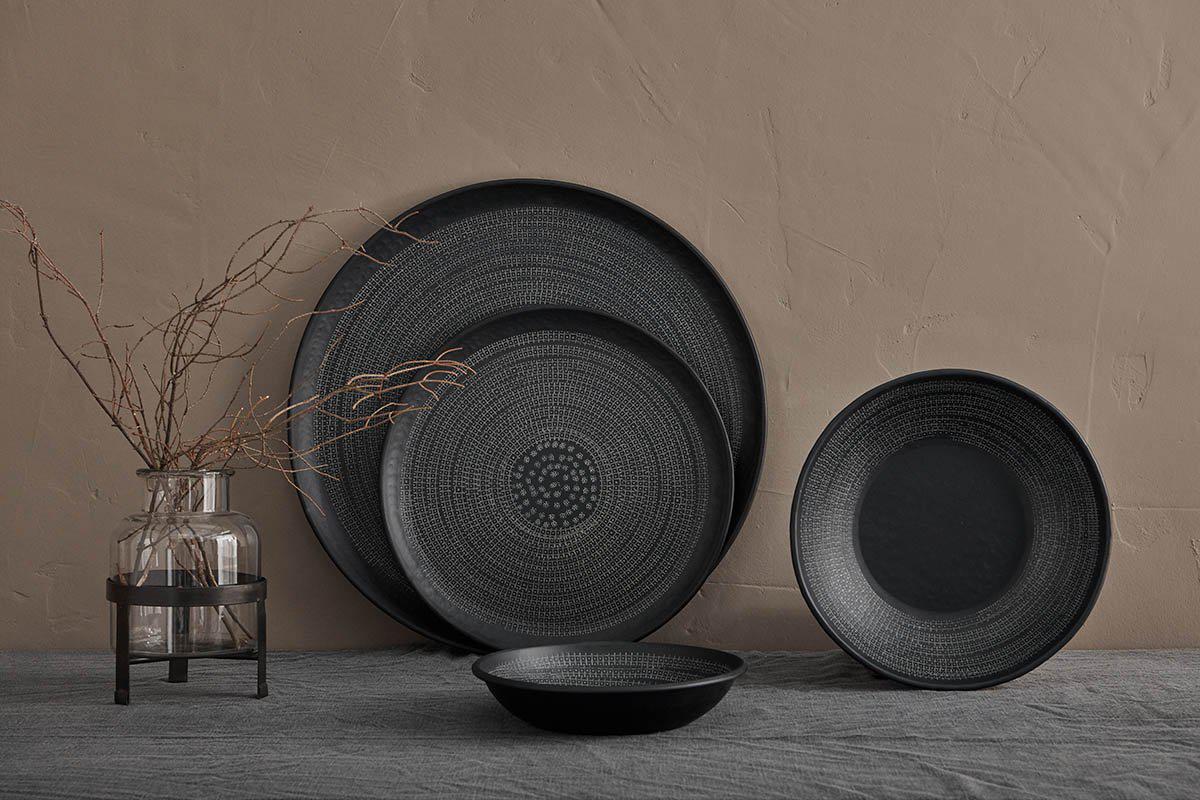 The nkuku Mahika Tray in black, featuring a dark, hand-etched ceramic texture, is styled on a gray fabric surface against a beige wall. A clear glass vase with bare branches completes this minimalistic and muted setting.