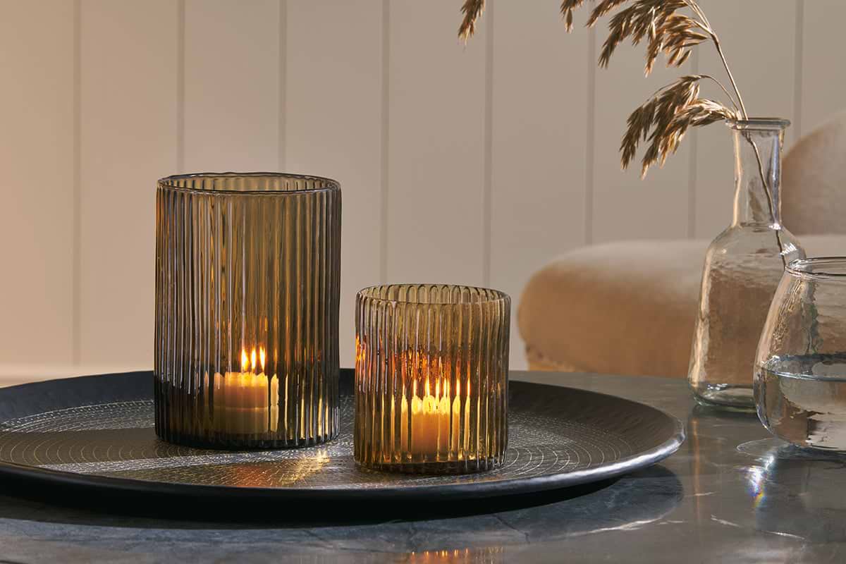 Two ribbed glass candle holders from nkuku—one tall and one short—sit on a round metallic tray, each holding a lit candle. The Malana Recycled Glass Candle Holder in smoke brown brings warmth and character to the arrangement. Nearby, a glass vase with dried grass and another with water complete the display, set against neutral-toned furniture and a paneled wall.