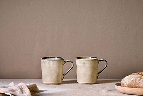 The nkuku Malia Mug - Cream (Set of 2) features textured rims and handles, styled side by side on a light surface with crusty bread and a folded cloth, set against a neutral wall for a minimalist, cozy vibe.