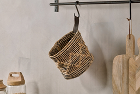 The nkuku Mannu Cotton & Hemp Wall Hung Basket in natural and black stripes hangs from a metal hook on a wall rod, with wooden cutting boards and a glass jar with a wooden lid nearby on a light surface against a neutral wall.