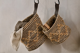 Two Mannu Cotton & Hemp Wall Hung Baskets by nkuku, featuring black and natural stripes, hang on a light wall. The left holds a beige cloth; the right has a wooden-handled brush. Both have dark leather handles for versatile storage.
