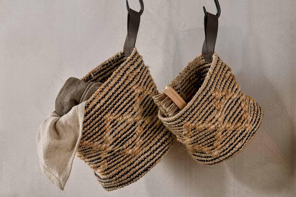 Two Mannu Cotton & Hemp Wall Hung Baskets by nkuku, featuring black and natural stripes, hang on a light wall. The left holds a beige cloth; the right has a wooden-handled brush. Both have dark leather handles for versatile storage.