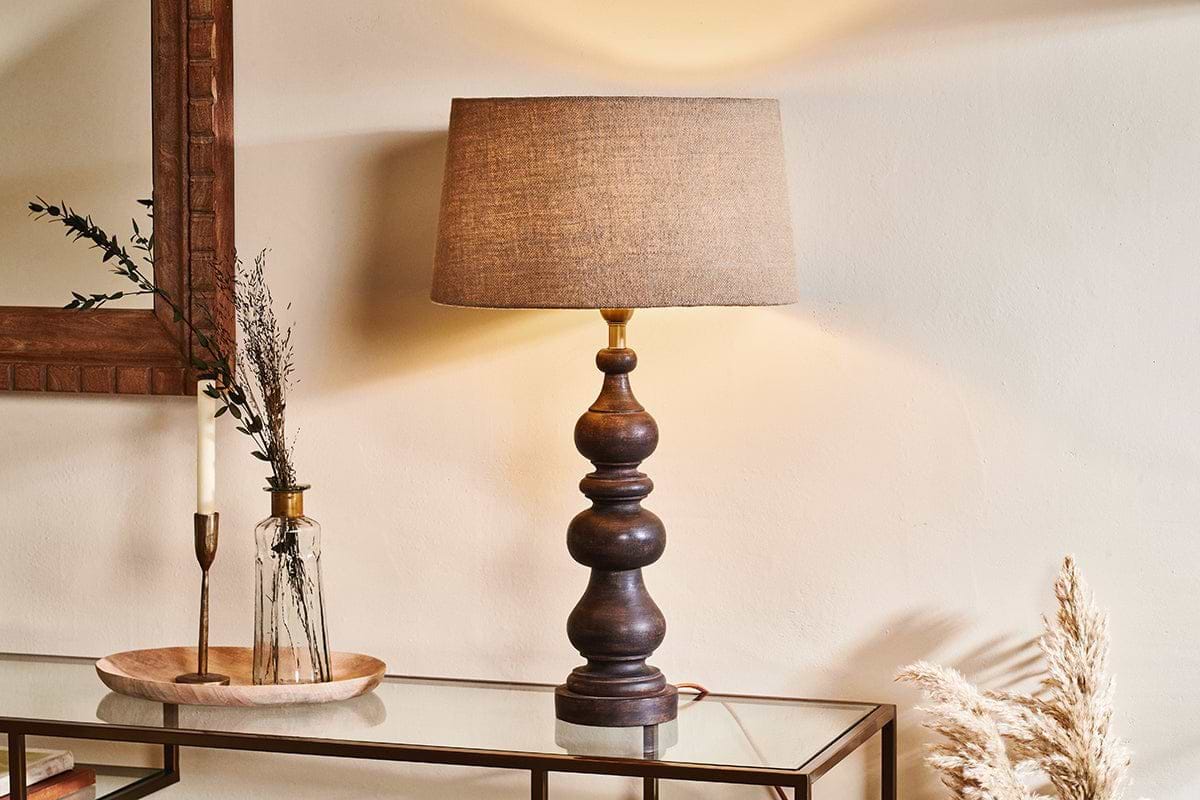 The nkuku Manua Wood Table Lamp - Large, featuring antique brass fittings and a beige fabric shade, sits on a glass console table with a clear vase of dried plants, candlestick, and wooden tray. Neutral walls and dried grasses complete the cozy decor.