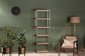 A minimalist room features the nkuku Marjori Mango Wood and Iron Standing Shelves - Narrow against a green wall, flanked by two potted plants, a black wall clock on the left, and a floor lamp with a wooden cane-back armchair on the right.