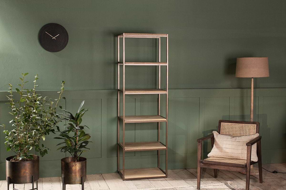 A minimalist room features the nkuku Marjori Mango Wood and Iron Standing Shelves - Narrow against a green wall, flanked by two potted plants, a black wall clock on the left, and a floor lamp with a wooden cane-back armchair on the right.