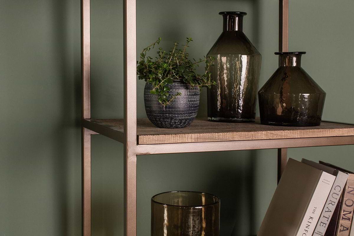 The Marjori Mango Wood And Iron Standing Shelves - Narrow by nkuku, set against a green wall, display a small leafy plant in a textured pot, two dark glass vases, a glass tumbler, and upright books for a minimalist modern look.