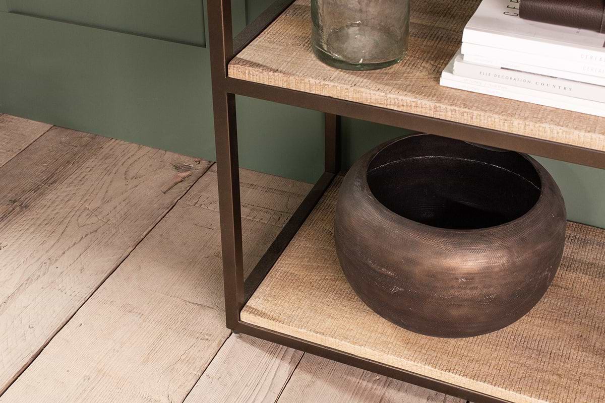 Close-up of the Marjori Mango Wood And Iron Standing Shelves - Narrow by nkuku, featuring a slim metal frame and mango wood. The lower shelf shows a dark brown bowl; the upper holds white books, a notebook, and clear glass against green wall and wooden floor.