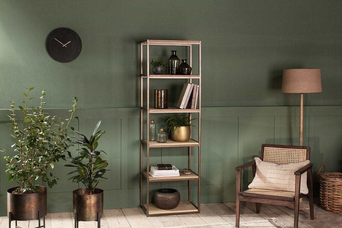 A cozy living room with a green wall, nkuku Marjori Mango Wood And Iron Standing Shelves - Narrow filled with books and decor, a black wall clock, two potted plants on stands, a wooden armchair with woven back, wicker basket, and floor lamp with beige shade.
