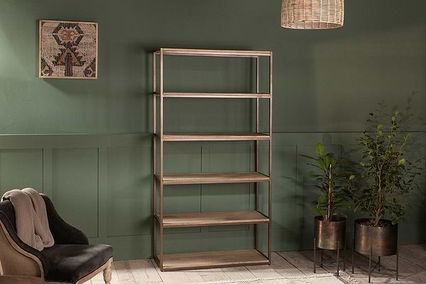 The Marjori Mango Wood And Iron Standing Shelves - Wide by nkuku stand empty against a green paneled wall, flanked by potted plants on black stands and a dark cushioned chair with a light throw. A woven pendant light and geometric artwork complete the look.