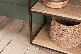 A close-up of nkuku's Marjori Mango Wood And Iron Standing Shelves - Wide displays a black metal frame with mango wood shelves. A round wooden bowl sits on the middle shelf and a large woven basket is on the shelf below, atop wide, weathered wooden planks.