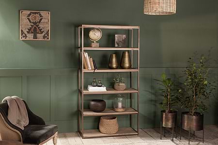 The nkuku Marjori Mango Wood And Iron Standing Shelves - Wide showcase books, vases, a globe, and plants against a green wall. Nearby potted plants, a plush gray chair, and a wicker pendant light create coziness beneath framed textile art.