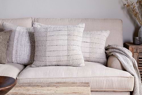 A beige sofa adorned with two Mayla Cushion Covers by nkuku—textured and cream-colored with subtle horizontal lines—sits elegantly in a living space defined by neutral tones. A chunky knit throw is draped over the arm, complemented by a rustic wooden coffee table in front and dried flowers in a vase on a wooden side table.