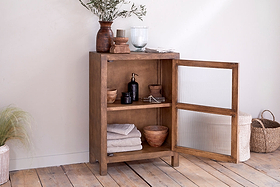 The nkuku Mendani Mango Wood & Fluted Glass Low Cabinet in Walnut Stain stands on a wooden floor, towels and soaps inside, with plants and dishes on top; woven baskets sit beside it against a light-colored wall.
