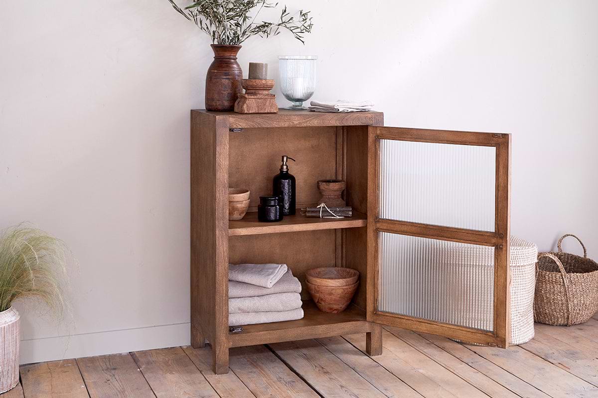 The nkuku Mendani Mango Wood & Fluted Glass Low Cabinet in Walnut Stain stands on a wooden floor, towels and soaps inside, with plants and dishes on top; woven baskets sit beside it against a light-colored wall.