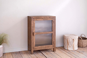 The Mendani Mango Wood & Fluted Glass Low Cabinet in Walnut Stain by nkuku stands on a light wooden floor, next to a lidded woven basket and an open handled basket. Ornamental grass decorates the corner against a white wall.
