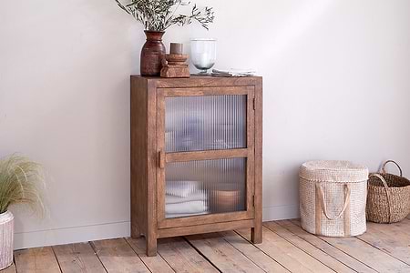 The Mendani Mango Wood & Fluted Glass Low Cabinet by nkuku in a walnut stain stands on a wooden floor, adorned with a vase, candle holder, and glass vase. Two woven baskets are positioned to its right.