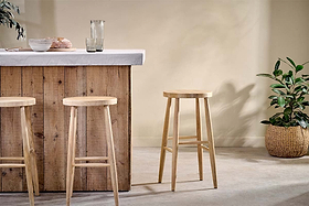 A rustic wooden bar with a concrete countertop features three nkuku Mengwi Mango Wood Round Bar Stools in natural. To the right sits a woven basket with a green plant, while a glass pitcher and two glasses are neatly arranged on the counter.