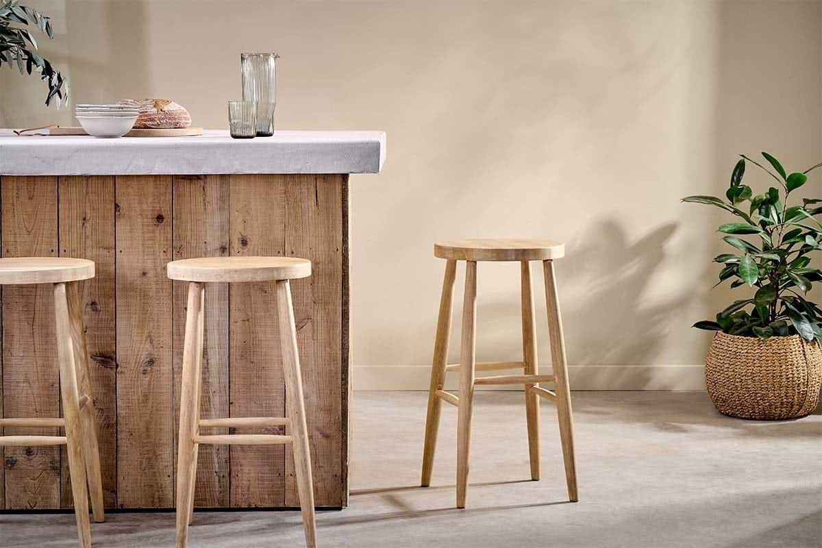 A rustic wooden bar with a concrete countertop features three nkuku Mengwi Mango Wood Round Bar Stools in natural. To the right sits a woven basket with a green plant, while a glass pitcher and two glasses are neatly arranged on the counter.