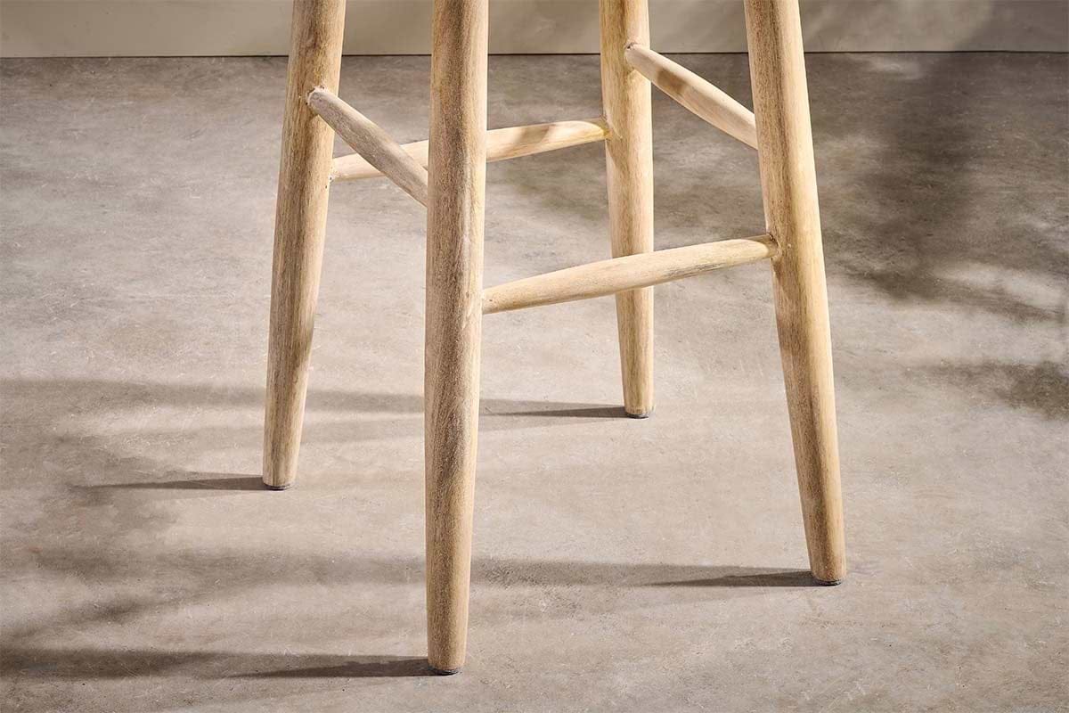 Close-up of the Mengwi Mango Wood Round Bar Stool by nkuku, showcasing its sustainable mango wood legs and rungs on a smooth, neutral-toned floor. Soft shadows play nearby, with a beige wall in the background.