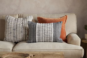 A beige sofa is adorned with two textured cushion covers—one square, white with black lines, and the other, the Mika Recycled Cushion Cover - Rectangle by nkuku, featuring black and white vertical stripes—beside a rust-colored velvet pillow. A wooden coffee table sits in front, adding warmth to the inviting home interior while a plant is partly visible on the right.