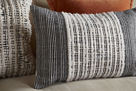 The Mika Recycled Cushion Cover - Rectangle by nkuku, featuring black-and-white textured stripes with small vertical dots, is displayed on a beige sofa. Behind it, a fuzzy cream pillow adorned with black lines and a solid rust-colored pillow complete the arrangement. Together, these stylish cushion covers create a cozy, layered look that elevates any home interior.